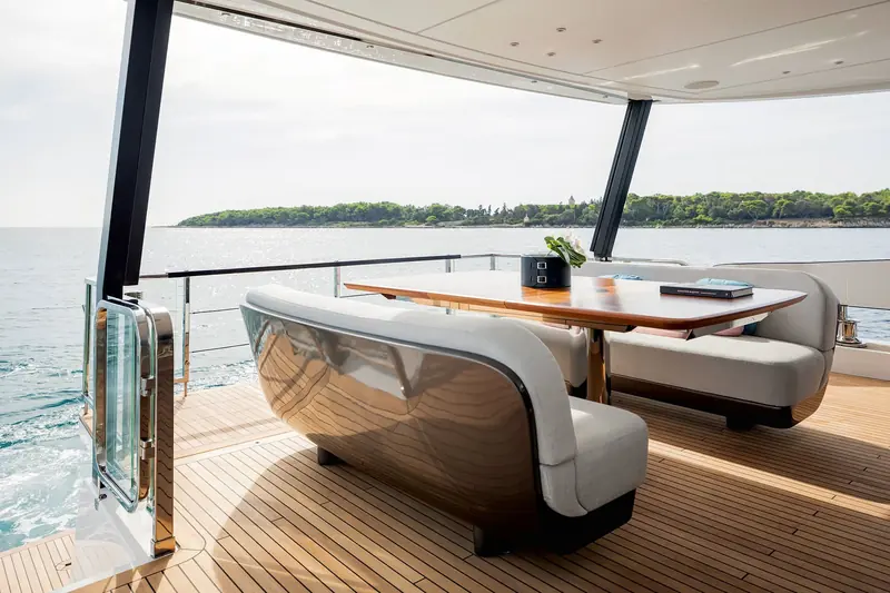 Aft cockpit dining under the hardtop, at anchor off a wooded Croatian island, with the polished stainless table leg and door furniture showing the finish level up close.