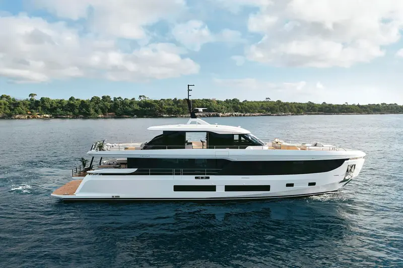 Azimut Grande 30M at anchor off a pine-covered headland, the full-beam main deck and low profile visible from abeam.