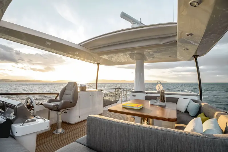 The open flybridge helm at last light, a second wheel and full instrument repeater set beside an L-shaped dinette under the hardtop roof.
