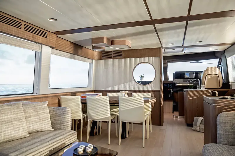 The saloon dining table set for eight, round portlight let into the bulkhead behind it and the galley visible through the open partition beyond.