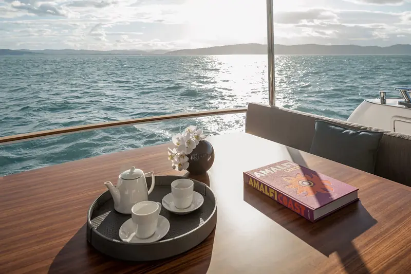 A coffee service laid out on the foredeck table at anchor, shells and a guidebook alongside it - the kind of quiet, shaded seating a full-beam foredeck lounge allows on a crossover hull.