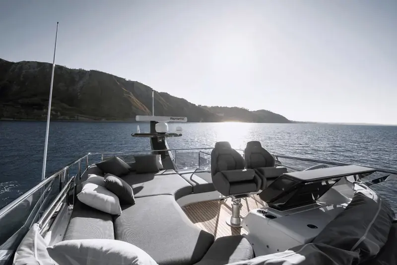 Looking aft from the sportfly deck at anchor near a rocky coastline, with the upper-station helm chairs, cushioned sunpad and folded bimini frame over the tender garage.