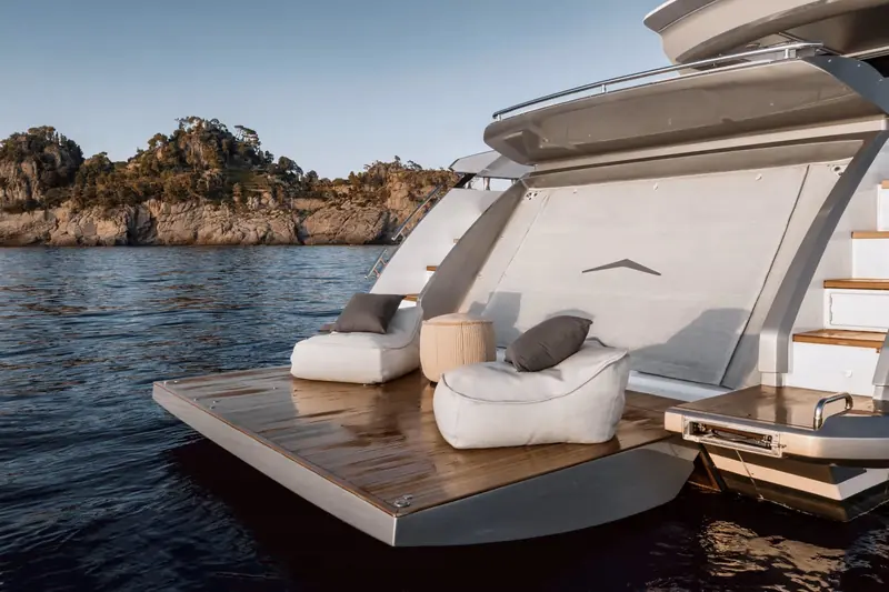 The fold-down transom platform lowered to the water off a rocky Riviera cove, beanbag loungers set out on the teak and the tender garage door visible above.