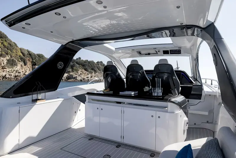 The three-across helm and companion seats under the hardtop, with the wraparound dash, drinks tray on the console and a rocky headland passing to port.