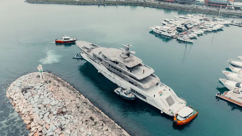 Tugs guide the newly floated Bilgin 243 past a marina crowded with moored yachts, her decks and windows not yet fitted out ahead of the outfitting phase.
