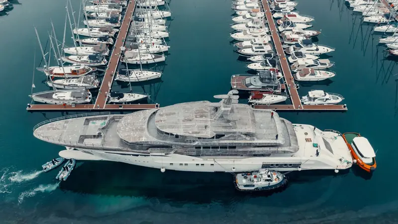 An overhead view of the Bilgin 243's bare hull threading between marina pontoons on tow lines, dwarfing the yachts moored on either side.