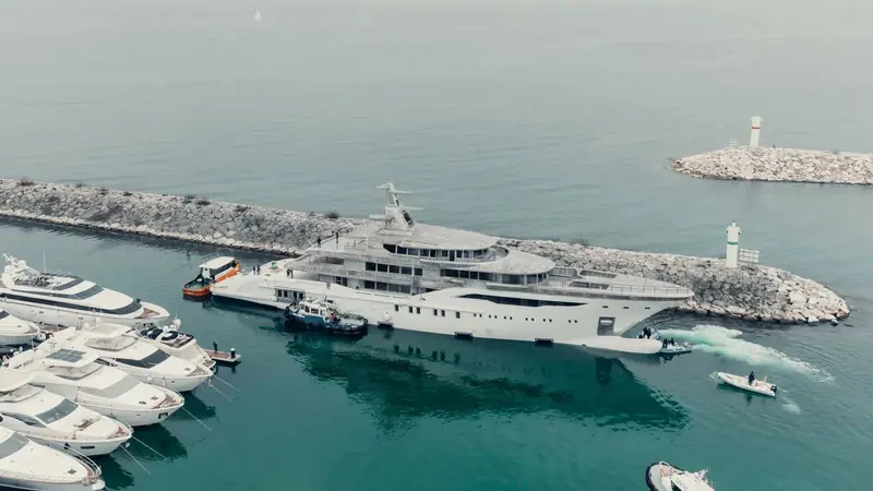 Two tugs walk the unpainted Bilgin 243 through the marina breakwater past a lighthouse, her raw hull mirrored in the flat water as onlookers on the transom watch the tow.