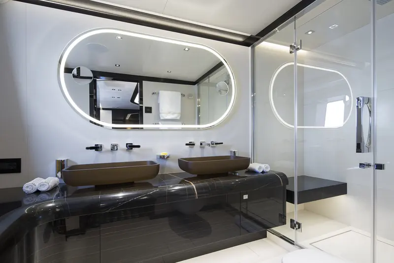 The full-beam ensuite bathroom off JAABER's guest suite, with backlit oval mirrors and a dark stone double vanity - Studio F.A. Porsche's material palette carried from the saloon into every head on board.