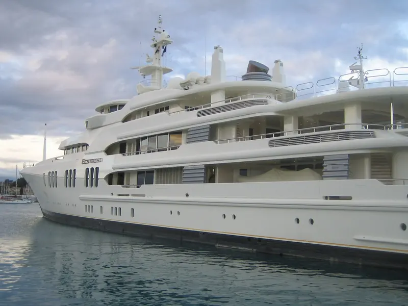 Bow-on at dusk in a Mediterranean marina, the row of oval hull windows lit from inside. Ecstasea's low, wide-shouldered profile was drawn to look purposeful rather than ornate, a look Feadship carried into several later builds.