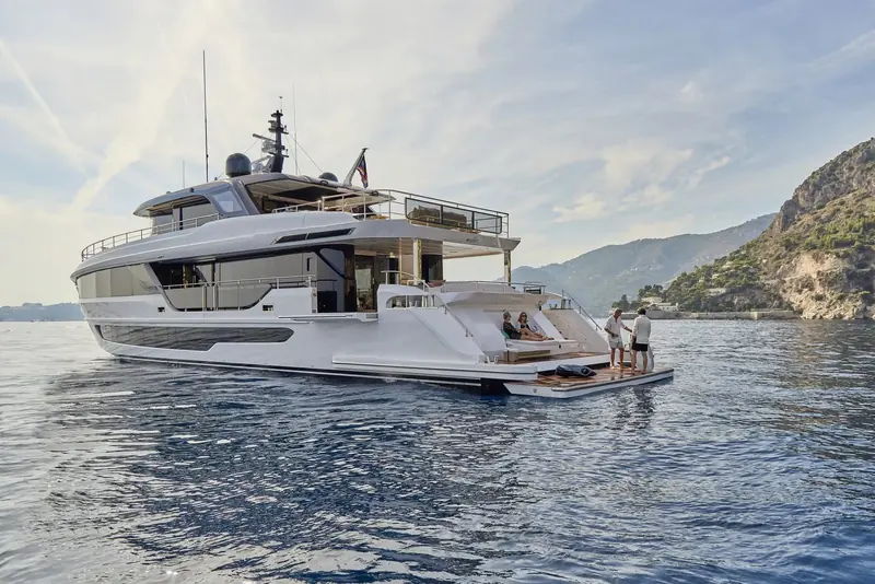 Two people on the swim platform off a Riviera headland, the tender garage door open above the transom and a rocky coastline behind.
