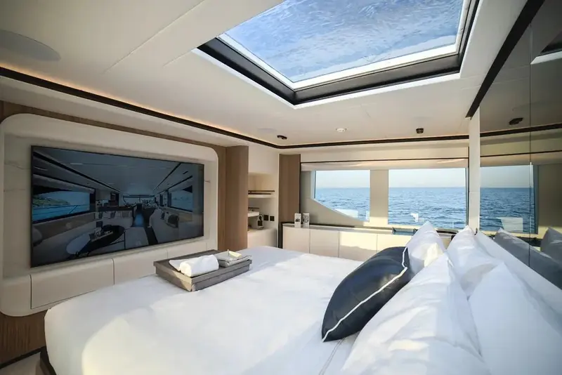 A guest stateroom lit by a full-length glass skylight over the berth, the headboard wall carrying an integrated wall-mounted screen.