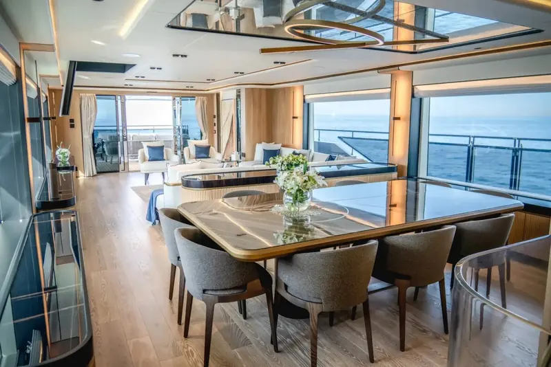 Main-deck dining table for eight set beneath a floor-to-ceiling window wall, the galley visible through the same open-plan run.