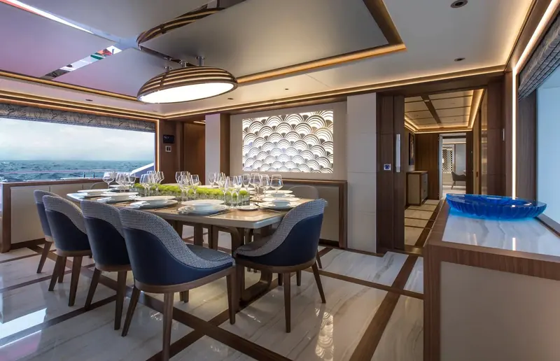A formal dining table for six sits inside the main-deck saloon, separated from the galley by a glass partition rather than a solid wall.
