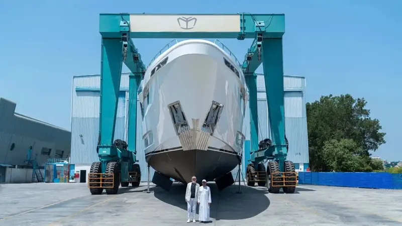Bow-on beneath the yard's twin-leg travel lift, the near-vertical stem and paired anchor pockets on full display as the shipyard principals pose for the launch photo.