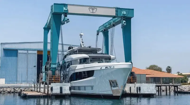 The grey-hulled sister ship lowers toward the water beside the yard's tile-roofed clubhouse, the crane's legs framing her superstructure.