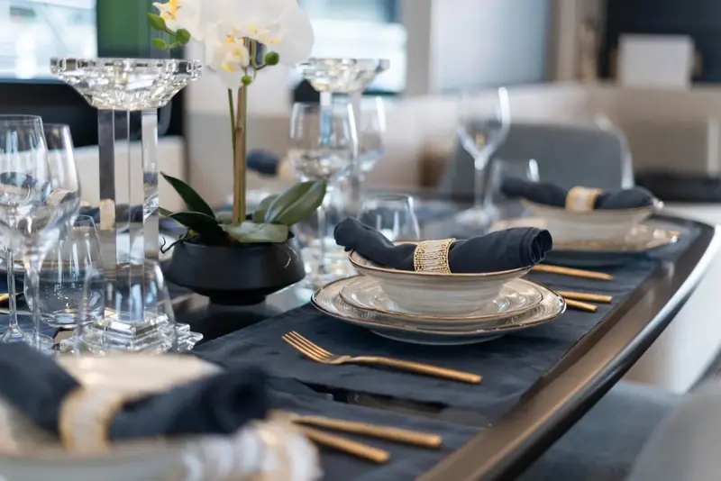 A formally laid dining setting on the same table, gold-rimmed china and folded navy napkins against dark wood.