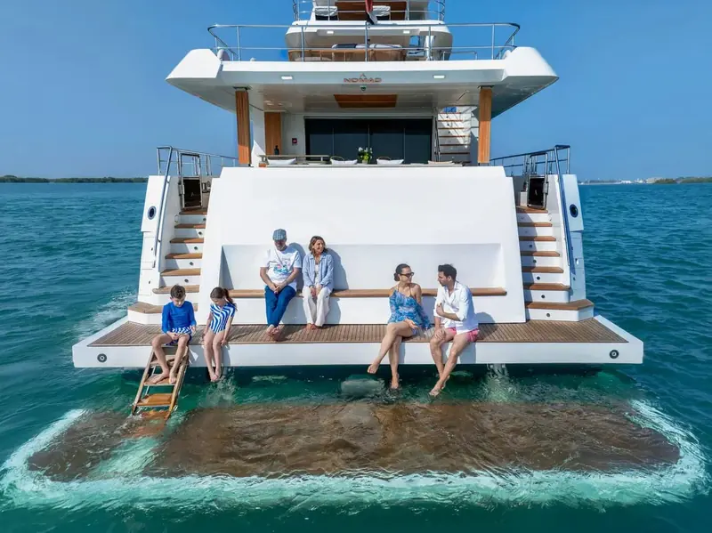 Guests wading off the fold-down swim platform at the stern, the boarding ladder deployed into shallow turquoise water.