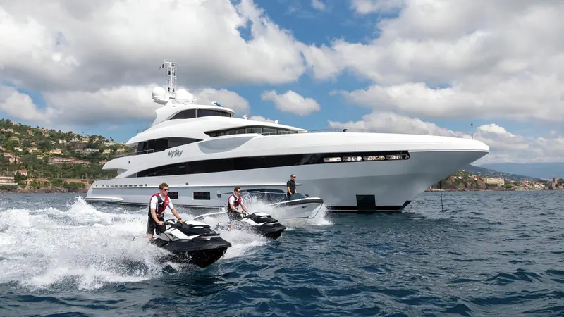 Guests ride jet skis alongside at anchor off a Mediterranean coastline, the tenders launched from a garage built into the transom rather than carried on deck.