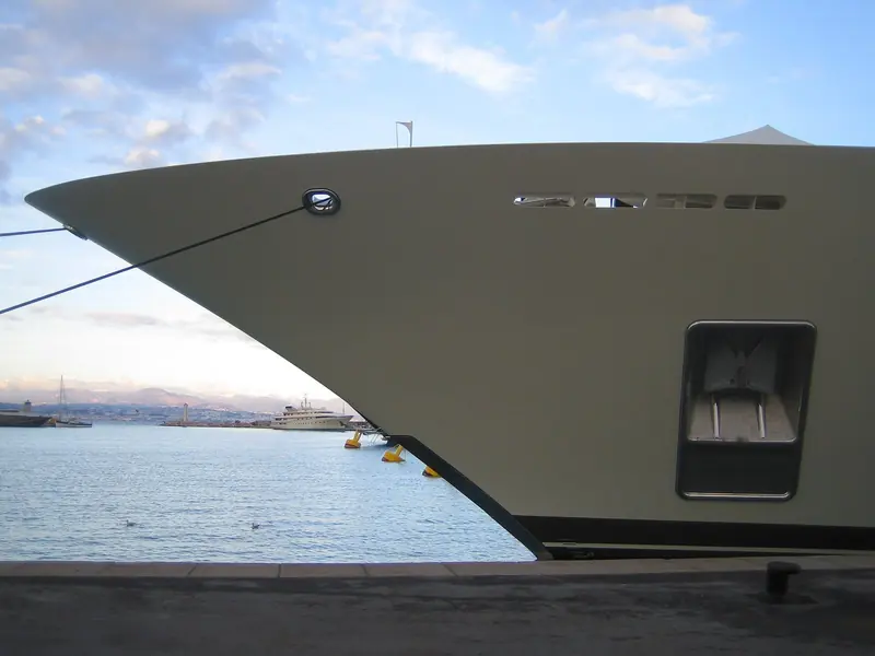 Dilbar's bow at anchor off a Mediterranean coastline, riding light with no crew visible on the exposed foredeck.
