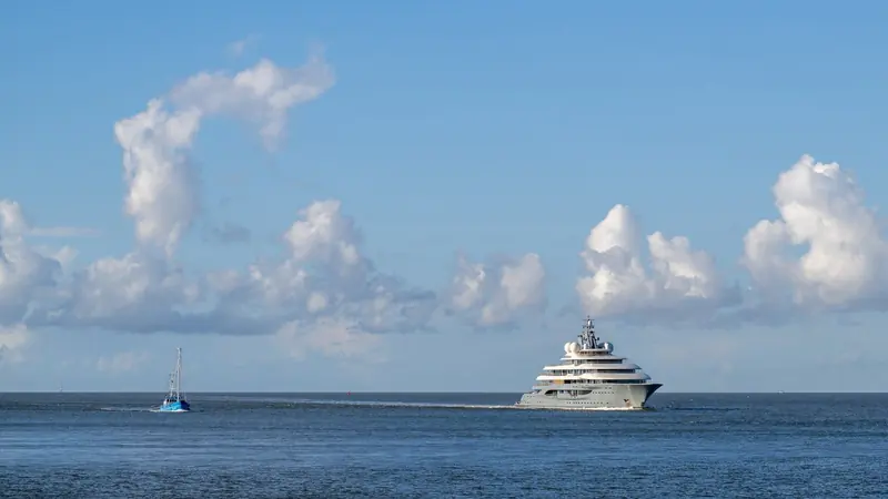 A distant profile against a bank of cloud, showing the near-vertical bow and low sheer line that mark Lurssen's later 130m-plus hulls.
