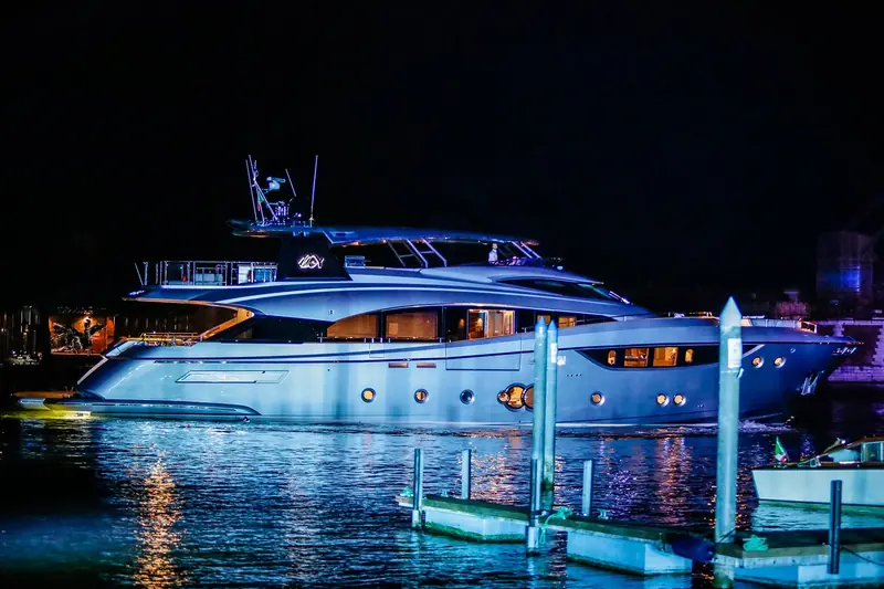 The first MCY 105 eases away from her Arsenale mooring poles at night, her hull washed in deep blue up-lighting between the wooden briccole.