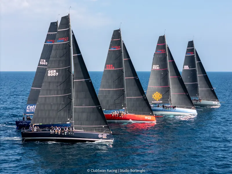 ClubSwan 50s at the start of a fleet race, sailing under matched black one-design mainsails.