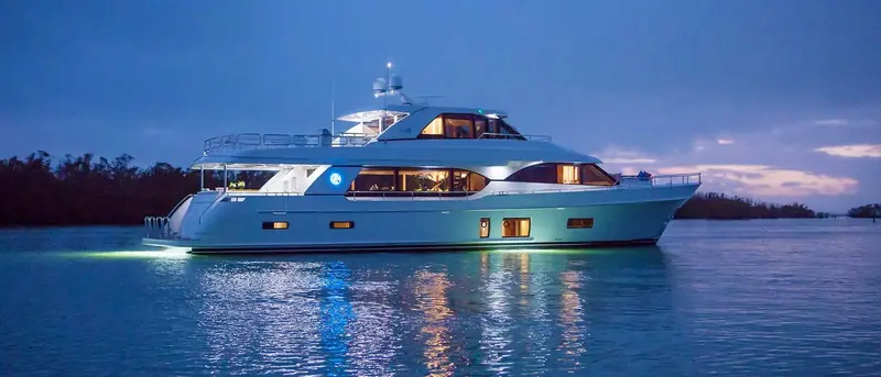 At anchor in blue-hour light, saloon and stateroom windows lit from inside, hull reflecting on still water.