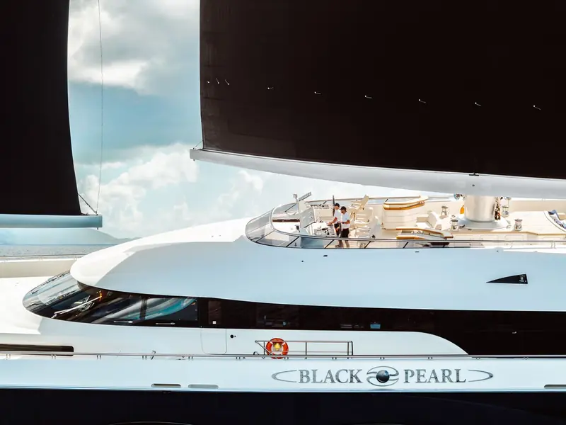 Crew on the aft deck beneath the black sails, with the yacht's name lettered across the transom in the style associated with her original owner, the late Oleg Burlakov.