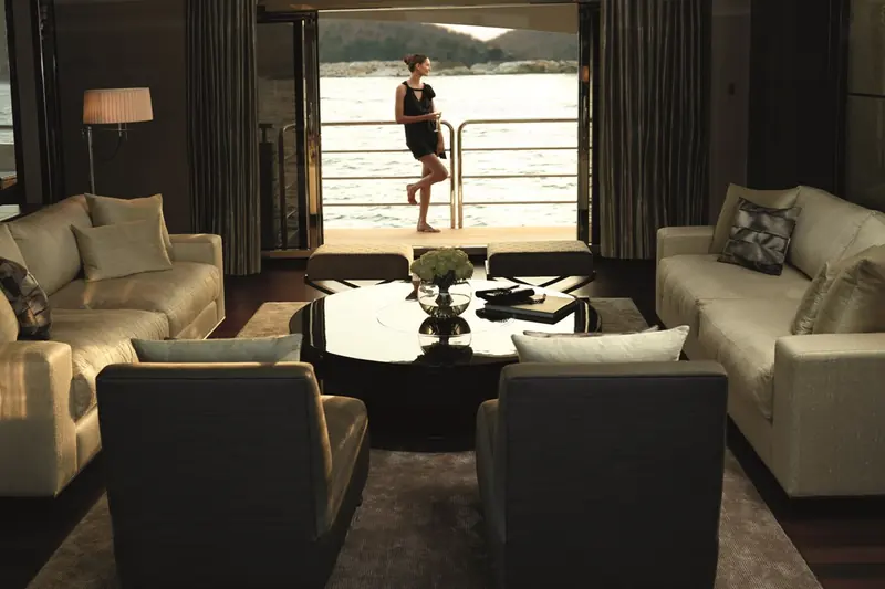 A guest steps out from the saloon onto the aft swim platform as the yacht sits at anchor off a wooded shoreline.