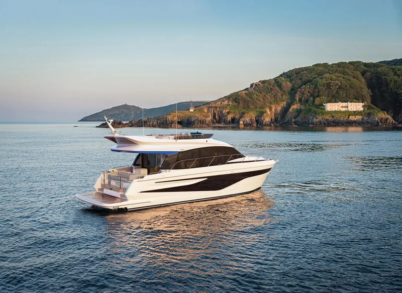 At rest in flat water off a wooded headland in low evening light. Seen side on, the flybridge sits low and well forward, and the hull window band runs almost the full length of the accommodation below.