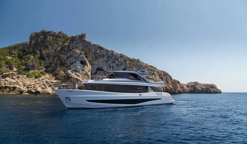 The X90 lying at anchor under a rocky headland topped by an old watchtower. The full profile shows the X Class signature: the superstructure is carried aft almost to the transom instead of stopping short as a conventional flybridge does, which is where the extra interior volume on this hull comes from.