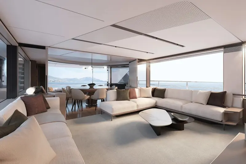The main saloon looking forward to the dining table and galley beyond. Glazing on both sides reaches down to seated eye level, so the coastline stays visible from the sofa, and the sole steps up only once between saloon and dining area.