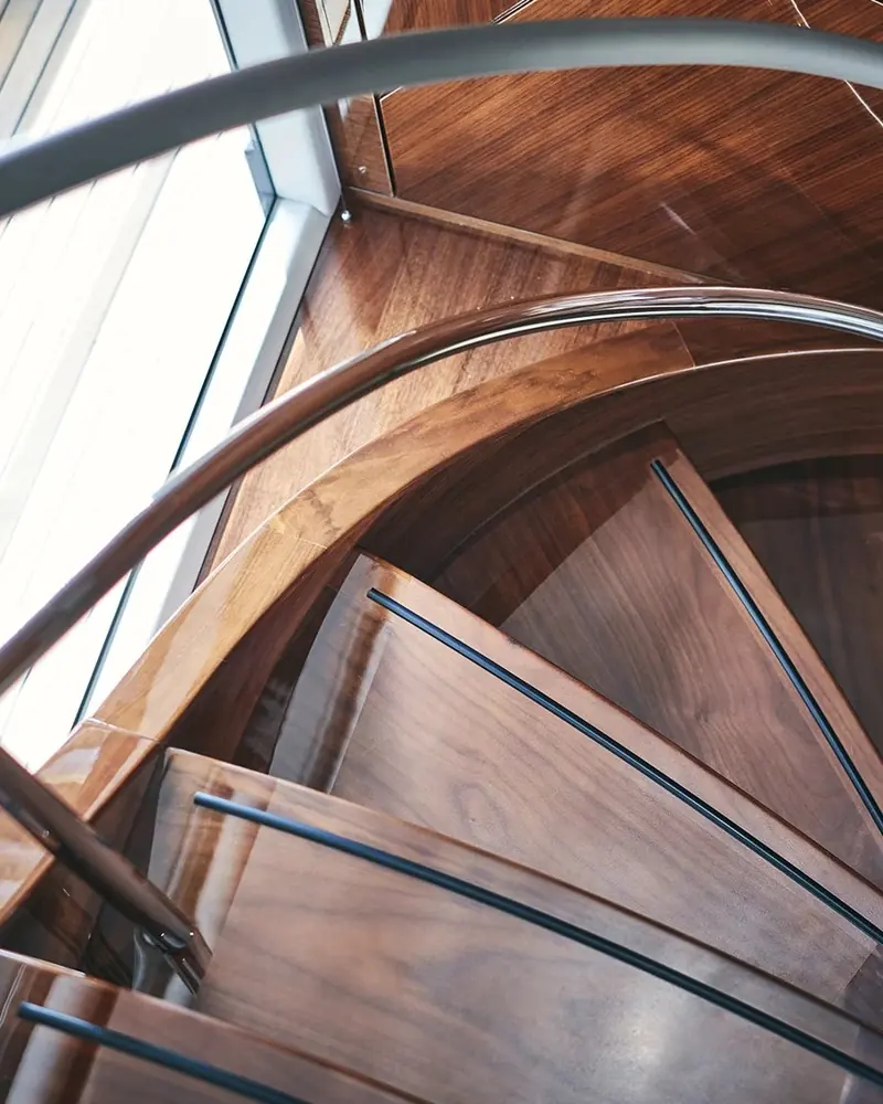 The walnut spiral stair seen from above. Each tread carries a dark inlaid stripe set back from its nosing, and the balustrade is a single curved glass panel rather than posts and wire, so nothing interrupts the view down the stairwell.