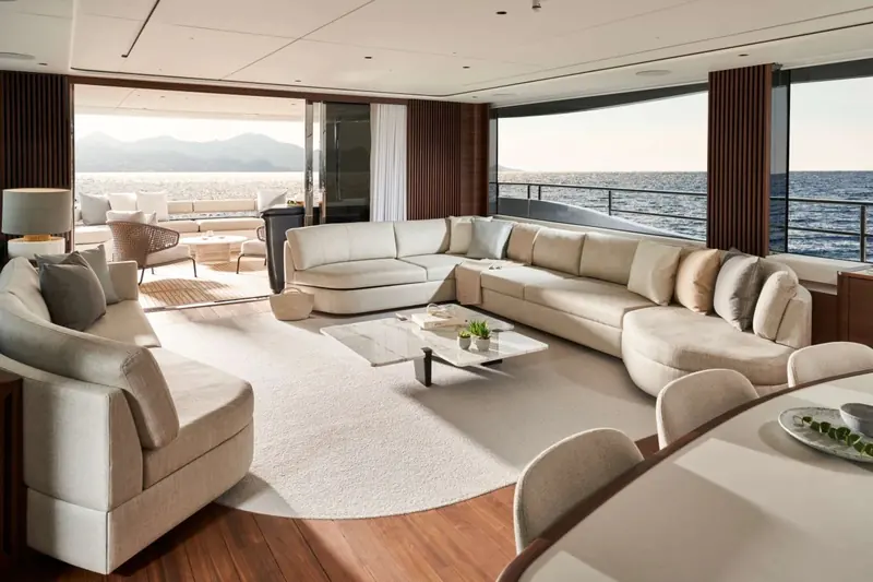 The main-deck saloon looking forward, walnut joinery against pale upholstery and floor-to-ceiling glazing on both sides.