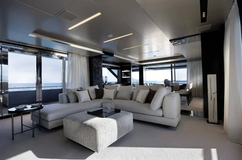 The main deck saloon opens to the sea through full-height glazing, with the dining table set forward of a low, wide sofa run.