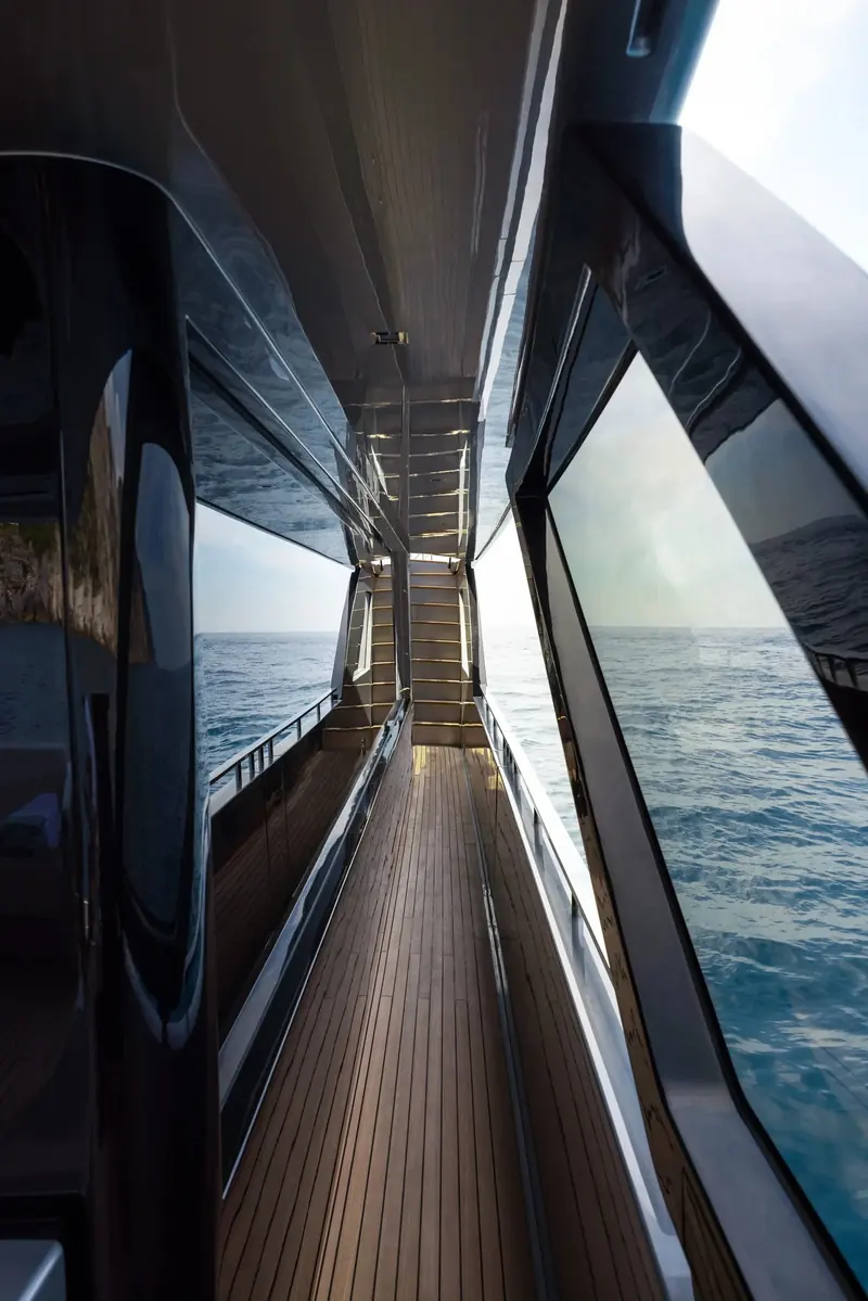 The starboard side deck runs glass-walled from bow to stern, throwing the reflection of the coastline back across the teak underfoot.