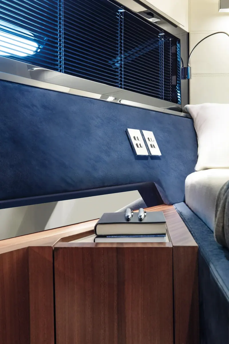 A cabin nightstand detail: venetian blinds over a hull window, USB outlets set into blue suede, and a mahogany-veneered cabinet, the marque's signature material mix below decks.