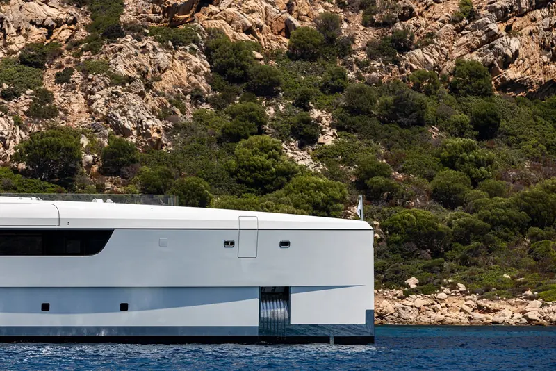 Cruising close along a rocky Mediterranean coastline, showing the low freeboard aft and the tender garage door built into the transom rather than carried on deck.