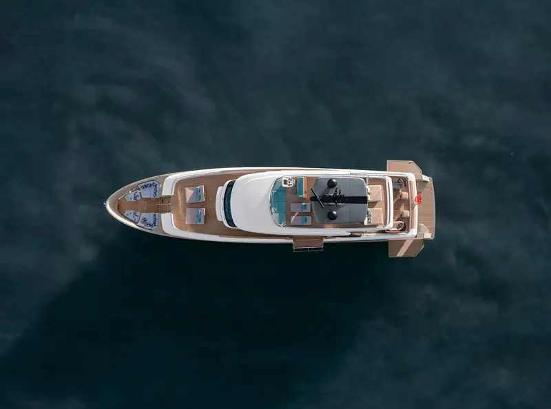A drone view straight down the foredeck shows the sunpad forward, the retractable helm-station glazing amidships and the shaded aft lounge, with teak decking running the full length.