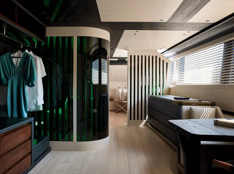 A cabin dressing area finished in pale oak and dark lacquer, with a curved glass-walled shower visible behind the wardrobe run.