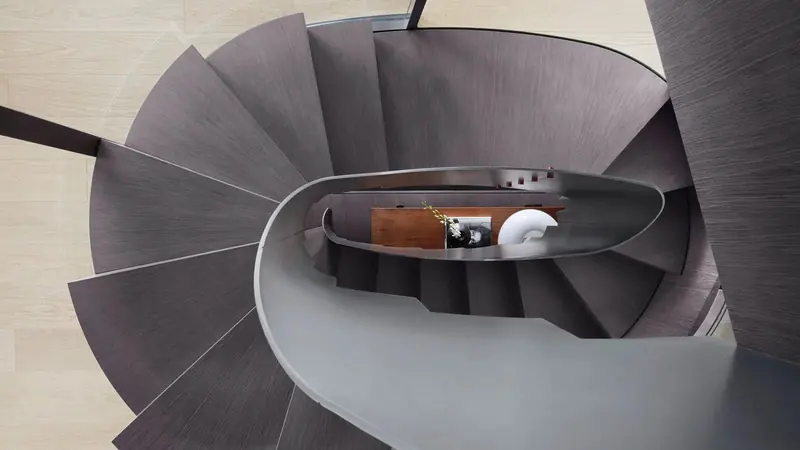 The glazed spiral stair connecting the decks, photographed from above: oak-veneer treads wrap a full turn around a landing where a stack of books and a framed print sit on a walnut console.