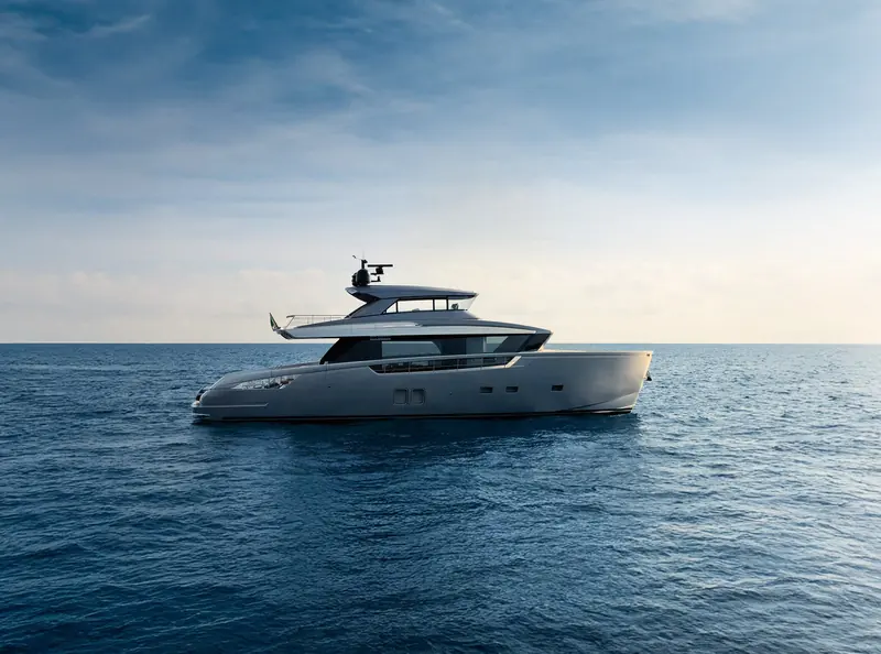 The Sanlorenzo SX76 under way in open water, her raised pilothouse and crossover hull line visible in profile with the Italian ensign flying aft.
