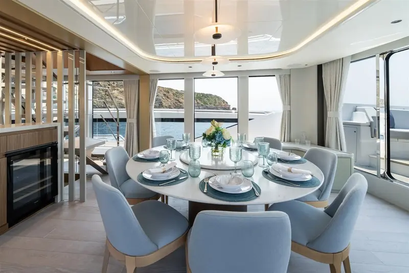 The dining table seats six under a glazed run of windows facing the anchorage, with the aft deck grill visible through the door beyond.