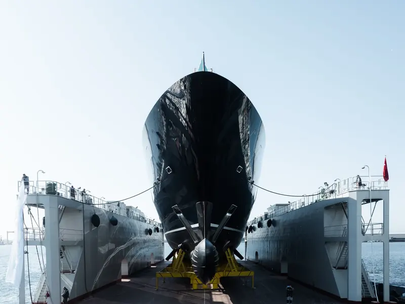 Bow-on in the yard's floating drydock before launch, twin propellers and shaft brackets still exposed, shipyard staff watching from the gantries either side.