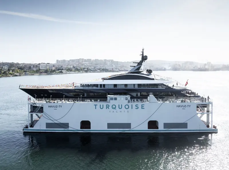 The yard's own transport dock, lettered HAVUZ-TY TUZLA and TURQUOISE YACHTS, towing the hull down the Bosphorus with crew and guests lining the rail.