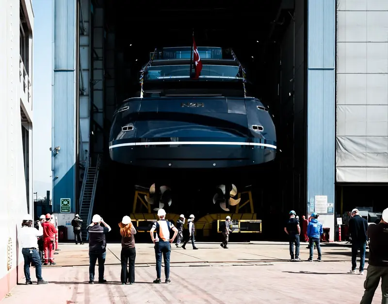 Shipyard workers and guests gather on the dock to watch the bow ease out of the covered launch hall, propellers still cradled on the transfer trolley.