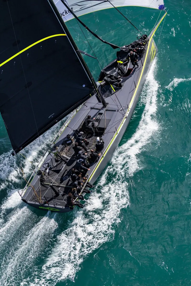 Close-hauled and heeled hard, crew hiking to windward as the yellow-striped hull slices through turquoise water.