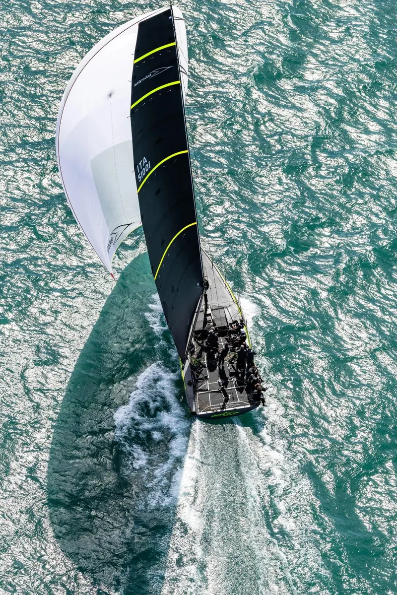 Overhead shot mid-tack with the crew crossing the deck and the mainsail luffing through the eye of the wind.