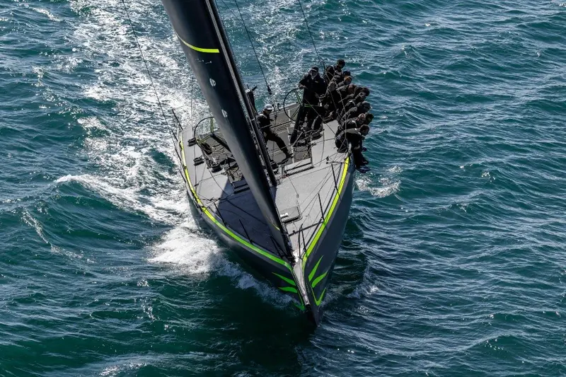 Powering upwind with crew stacked on the weather rail, the black carbon hull throwing a clean, narrow wake.