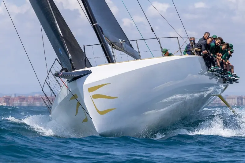 Bow-on in a header, the hull's gold flame graphic breaking the surface and the crew braced along the windward rail as spray sheets off the topsides.